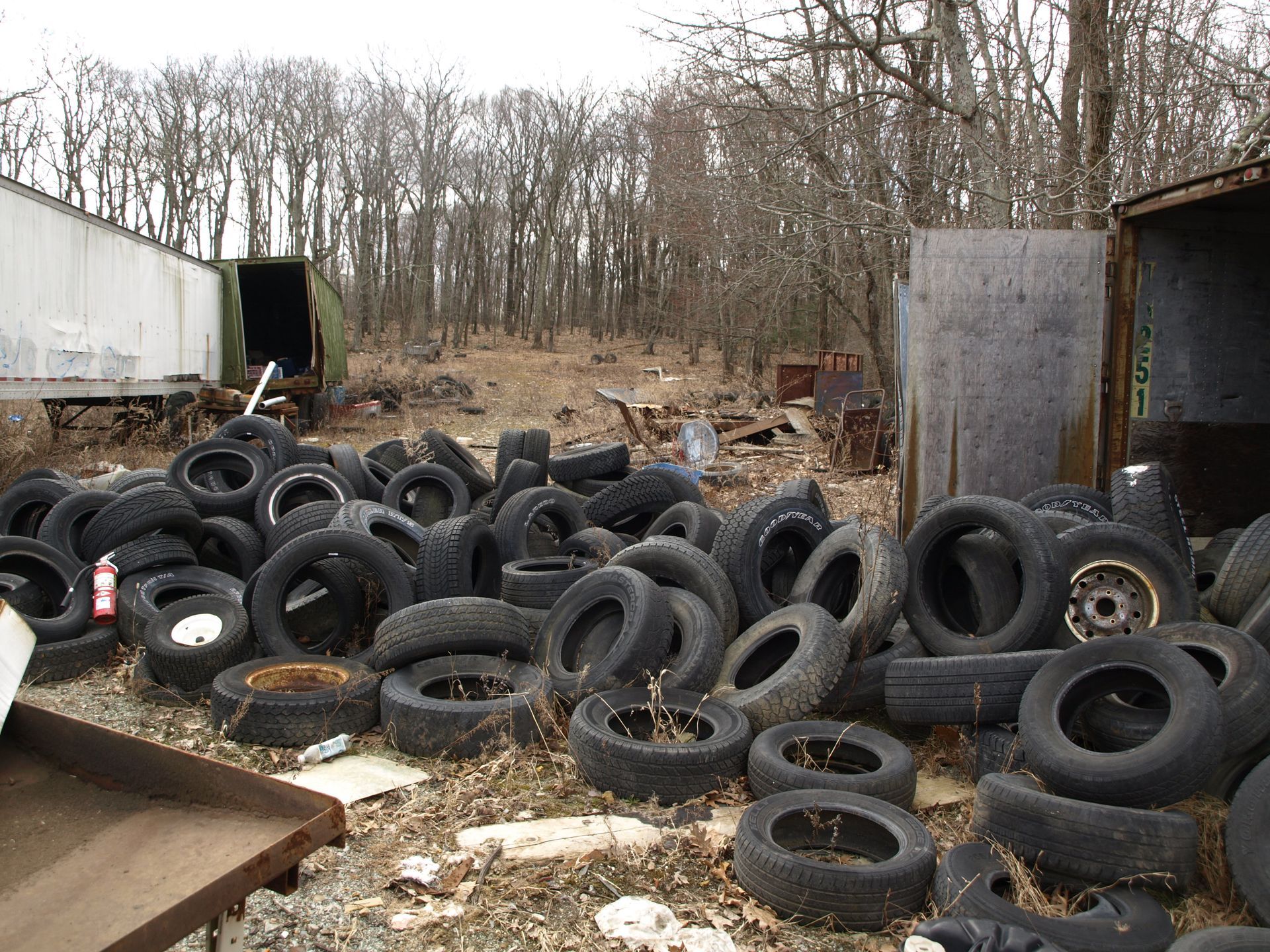 Pile of tires and debris in a wooded area, with trailers in the background.
