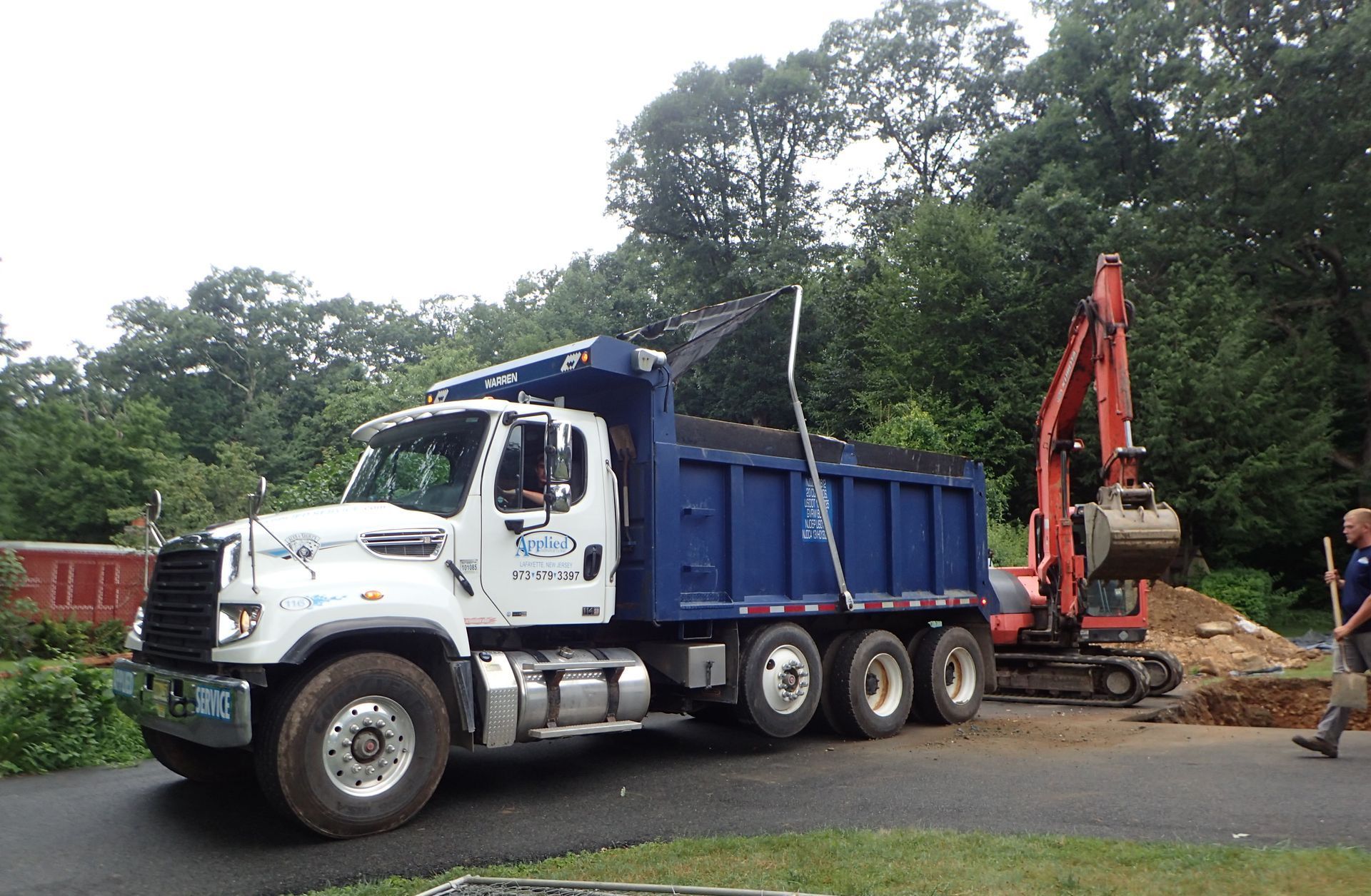 Dump truck and excavator at a construction site, blue truck, digging hole, and worker present.