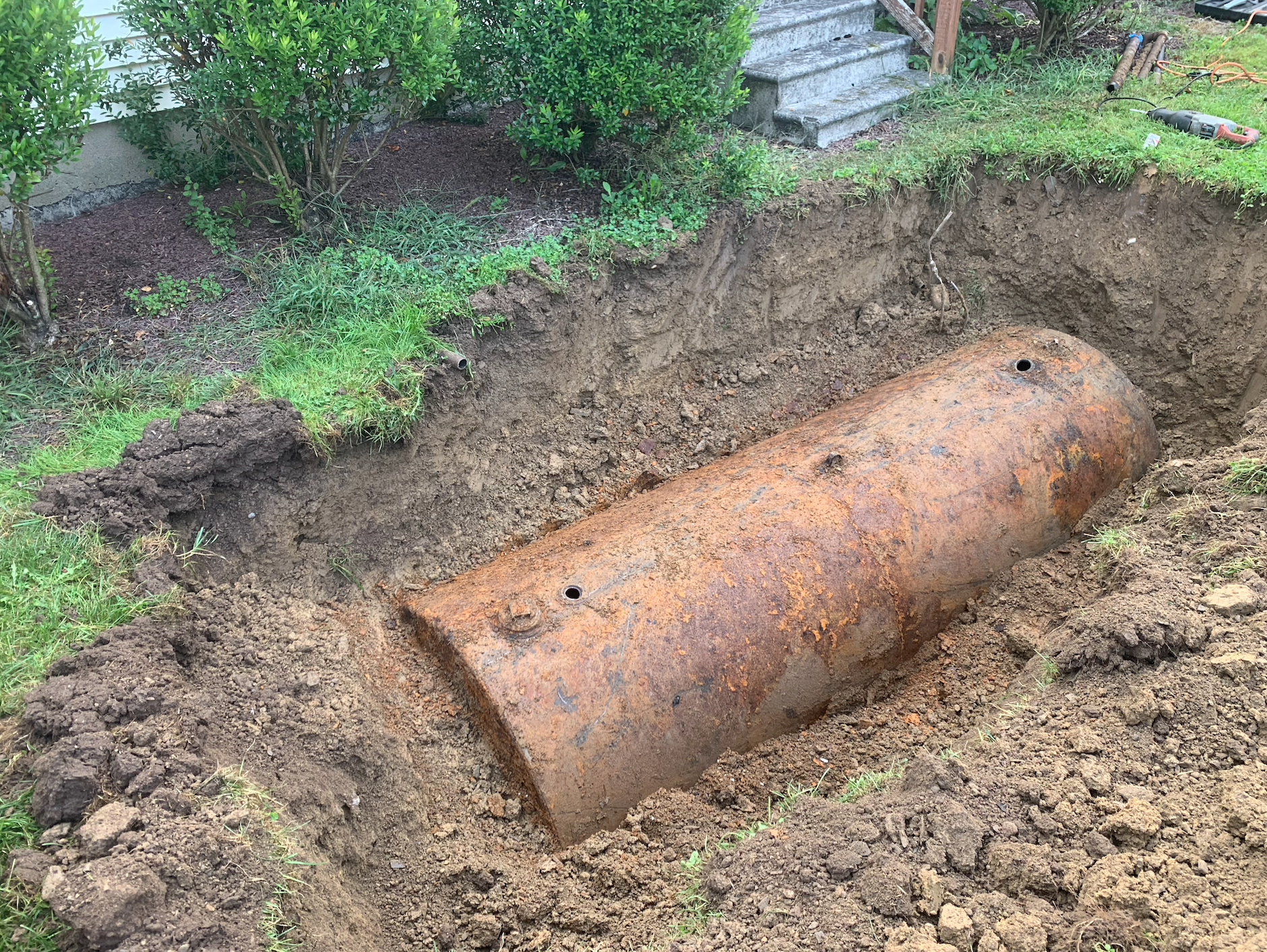 Rusty, buried metal tank in a dug-out pit; in a grassy yard, near bushes and steps.