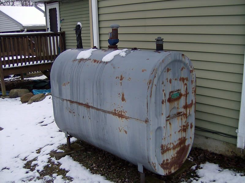 Exterior oil tank, weathered gray with rust, in a snowy yard next to a building.