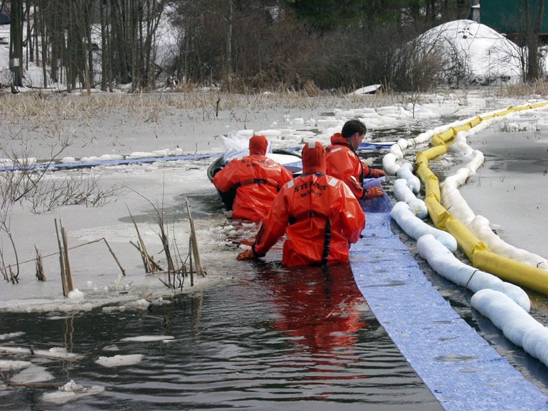 Workers in orange suits deploying a containment boom in icy water, presumably for an oil spill cleanup.