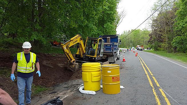 Roadside scene: Yellow excavator and dump truck, hazardous waste barrels, worker in safety vest, clearing debris.