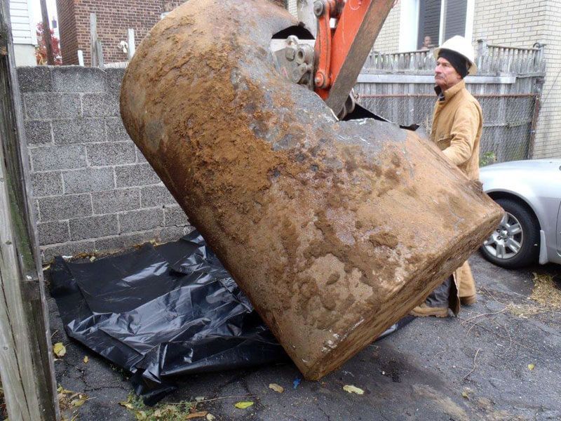 A worker in coveralls and a hard hat with an excavator lifting a rusty oil tank.