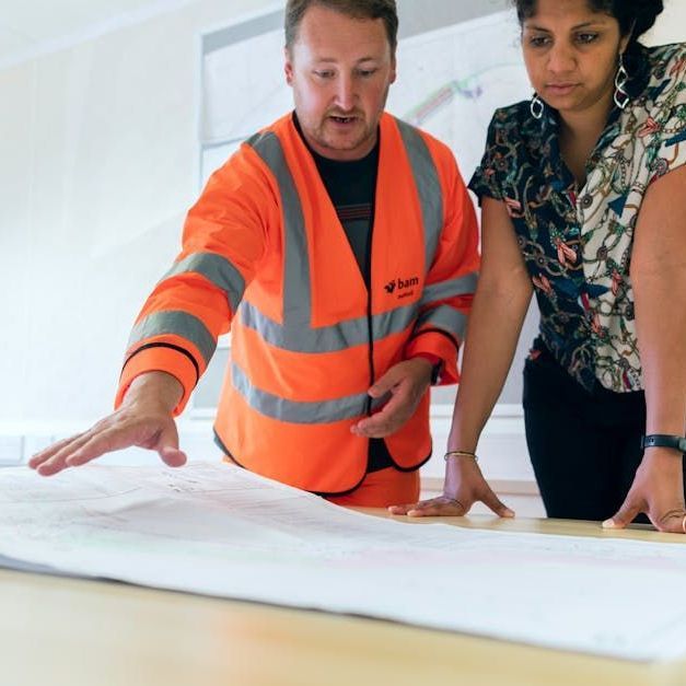 Man in orange safety vest and woman looking at blueprints, discussing plans.