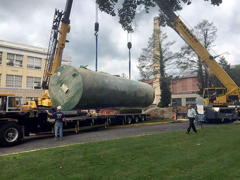 Two cranes lift a large, cylindrical tank above a flatbed truck in a grassy area near buildings and trees.