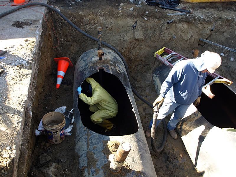 Workers inspect an underground tank. One wears protective gear inside the tank. Another uses a hose.