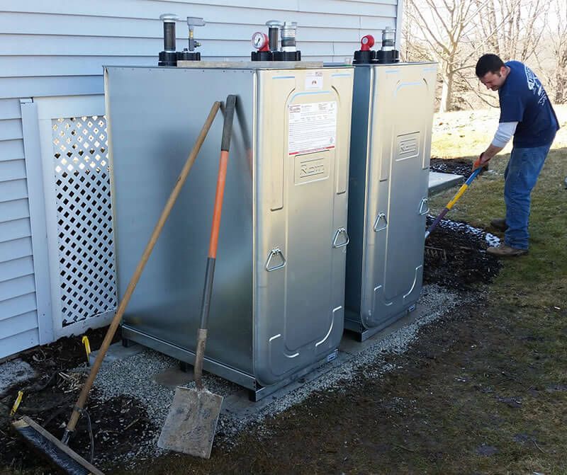 Man shovels gravel next to two metal tanks installed near a white building. Shovel leans against the tanks.