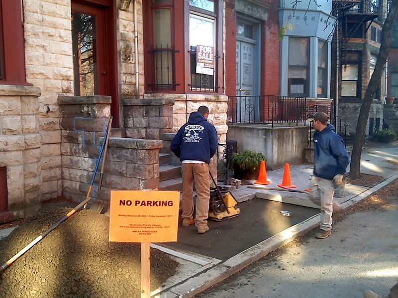 Two construction workers compacting asphalt on a sidewalk in front of brick buildings, 