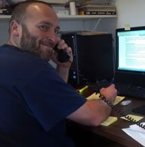 Man with beard, smiling, on the phone, writing notes at a desk with a computer and sticky notes.