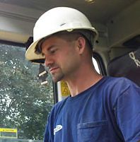 Man in blue shirt and white hard hat, looking to the left. In a vehicle.