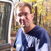Man with glasses smiling, standing by a white vehicle in a wooded area with fall foliage.