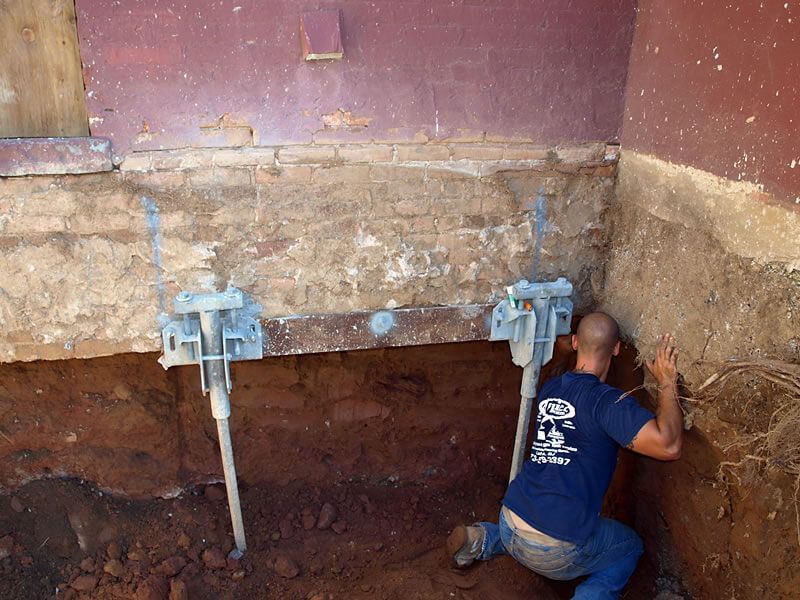 Man installing foundation supports on a brick building corner. Brown dirt and walls, silver jacks.