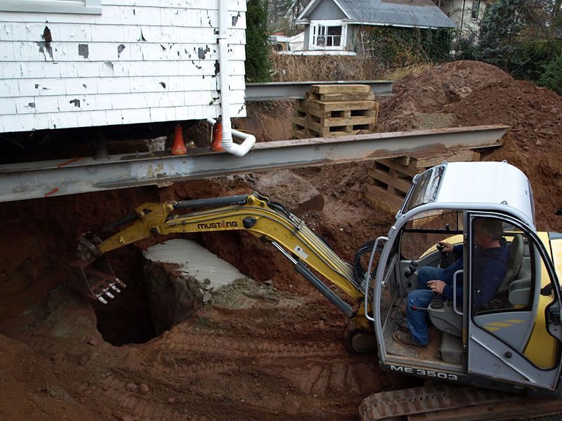 Construction site. Excavator digging near a house being lifted. A worker is in the machine. Brown dirt.