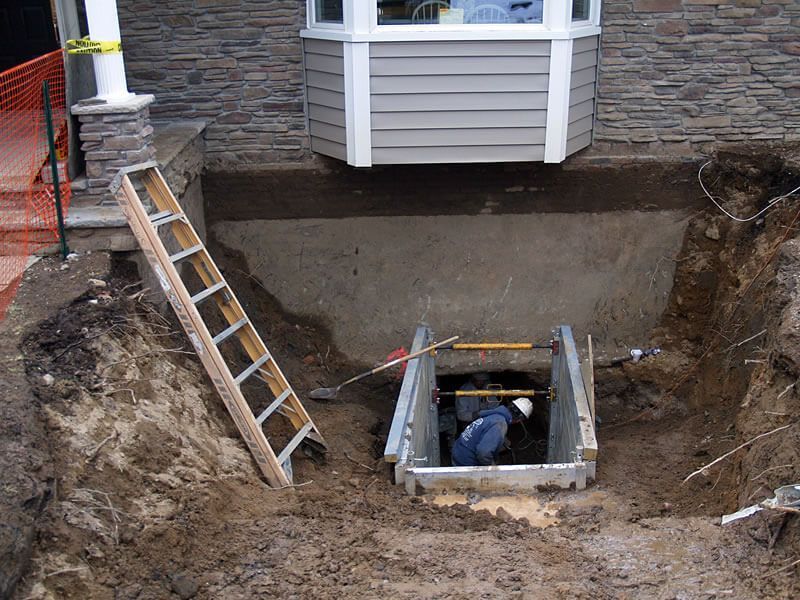 Construction site: worker in trench with metal supports, ladder, and soil excavation near a house.