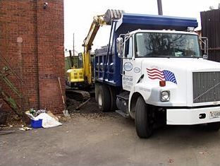 A backhoe loading debris into a blue dump truck beside a brick building.