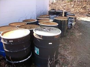 Group of black barrels with lids outdoors, near a building.
