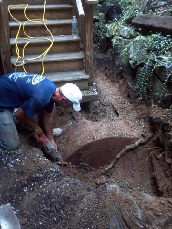 Man in blue shirt and white hat cutting a buried rusty metal tank near wooden stairs.