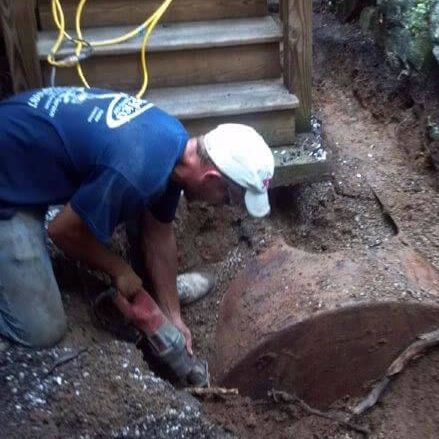 Man kneeling, using a saw to cut into a rusted metal tank in a dirt trench. Steps in the background.