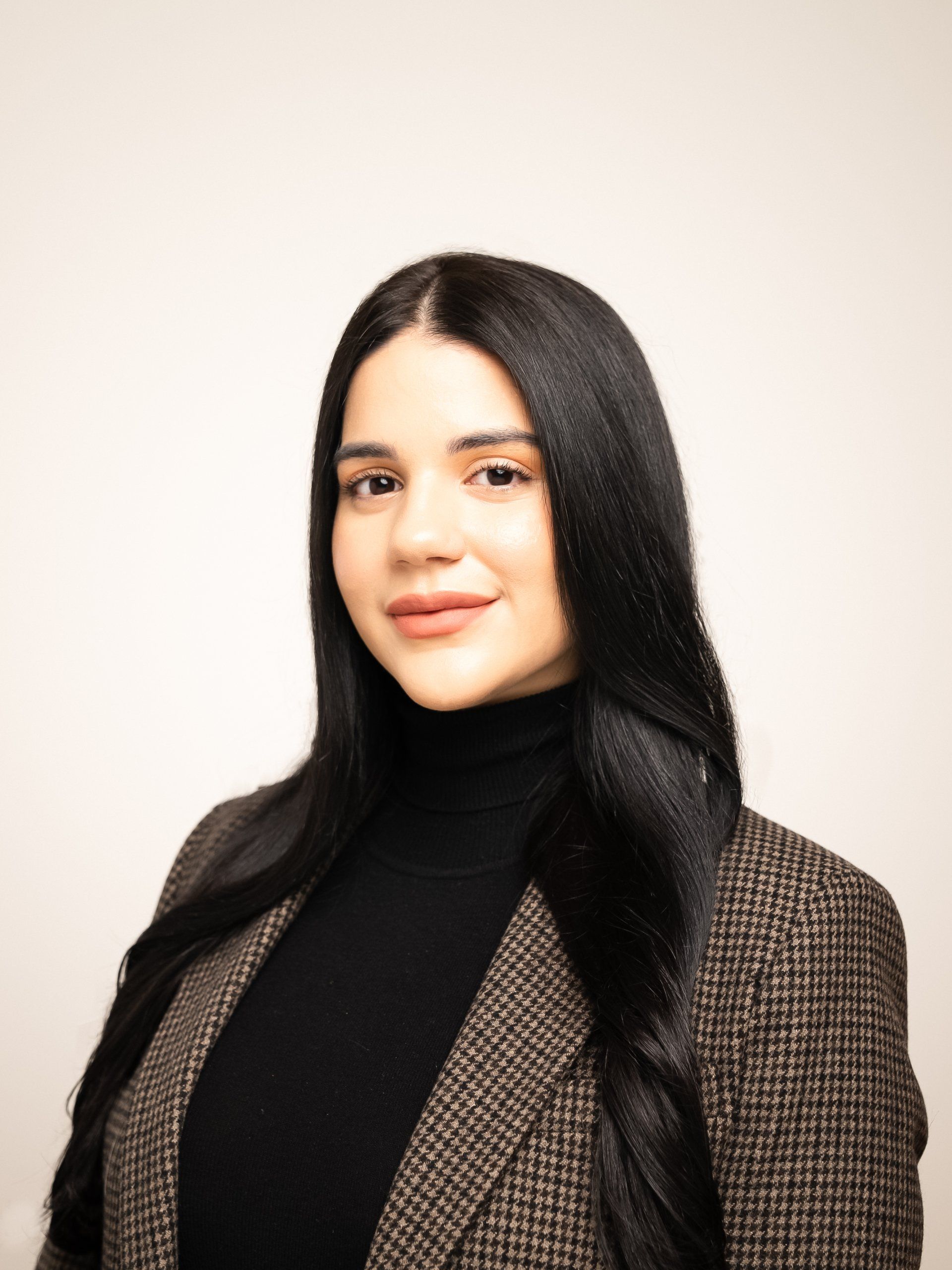 A woman with long black hair is wearing a plaid jacket and a black turtleneck.