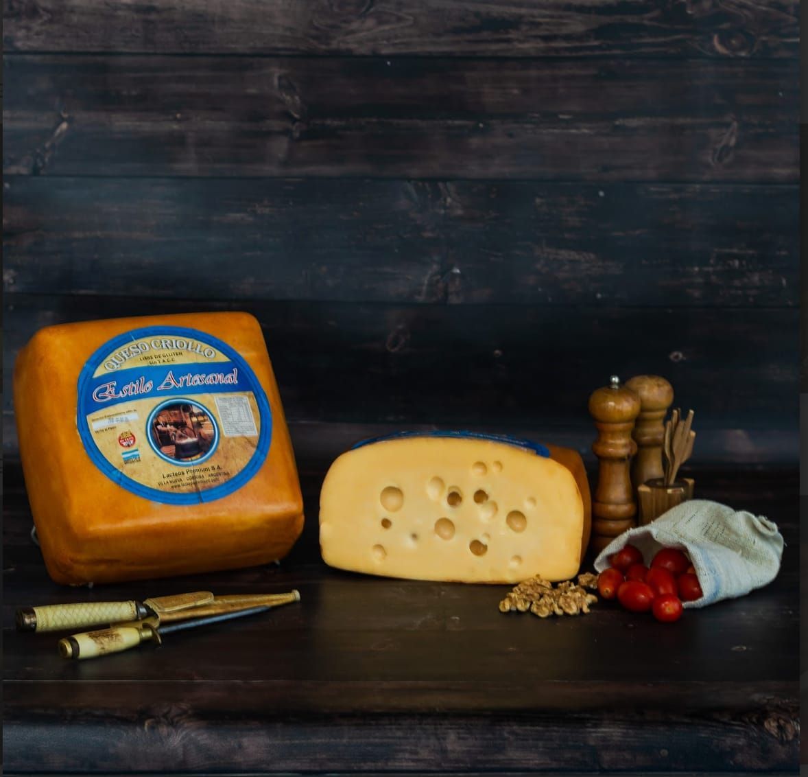 Bloque y cuña de queso con tomates, nueces y saleros y pimenteros sobre superficie de madera oscura.
