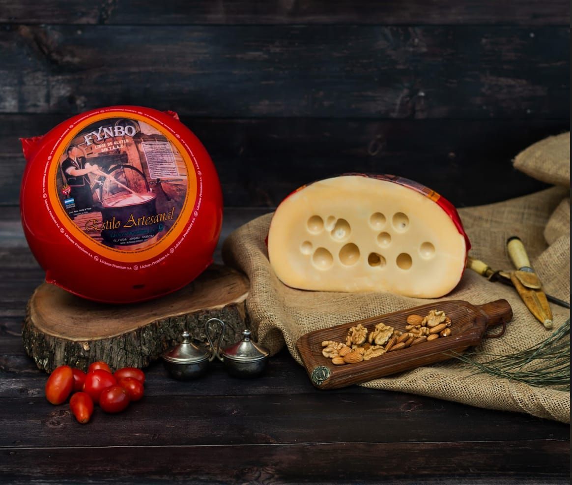 Queso entero redondo, encerado de color rojo y en cuña, con agujeros, sobre madera, arpillera y tomates.