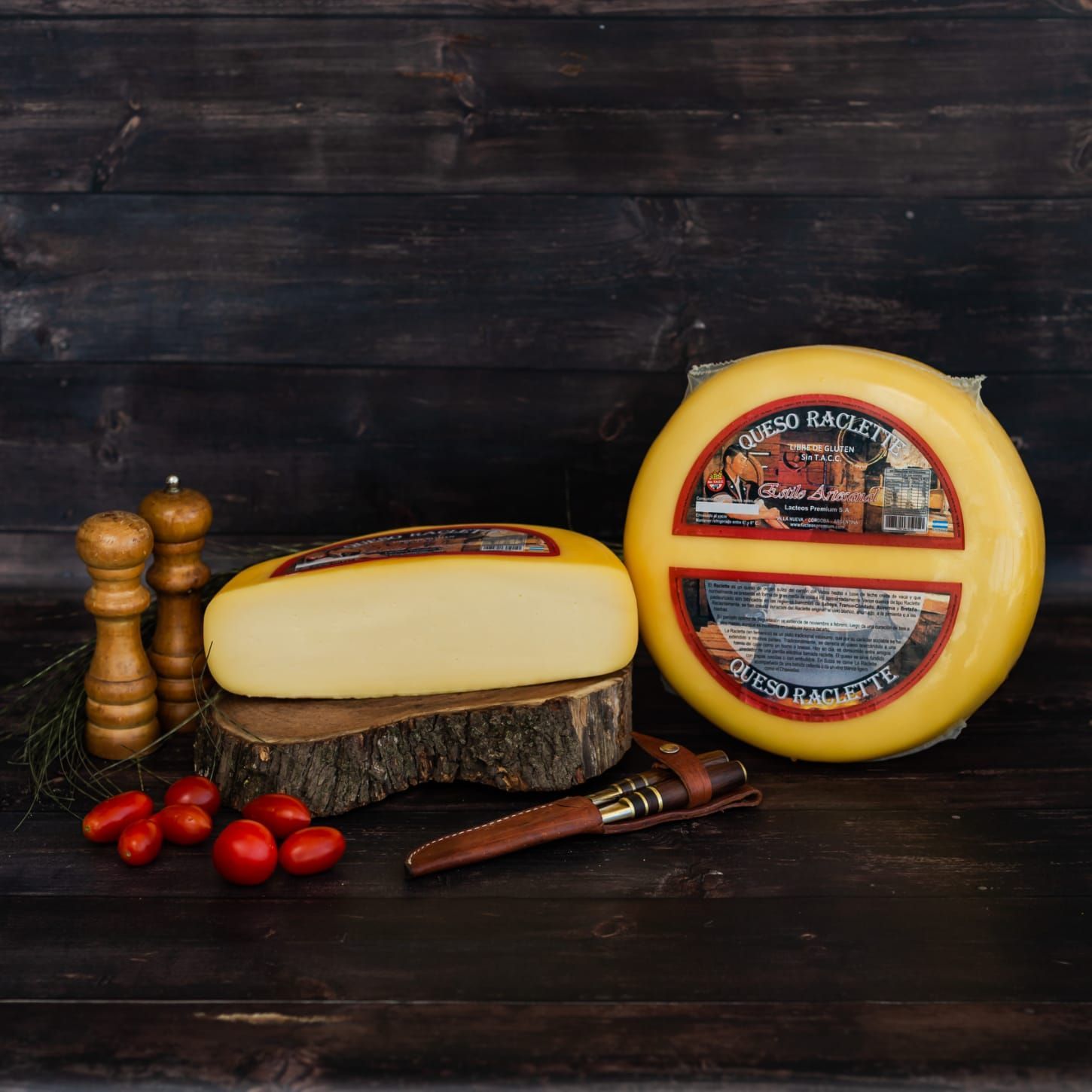 Redondo de queso con corteza amarilla, cortado en gajos sobre tabla de madera, tomates, molinillos de pimienta y cuchillo.
