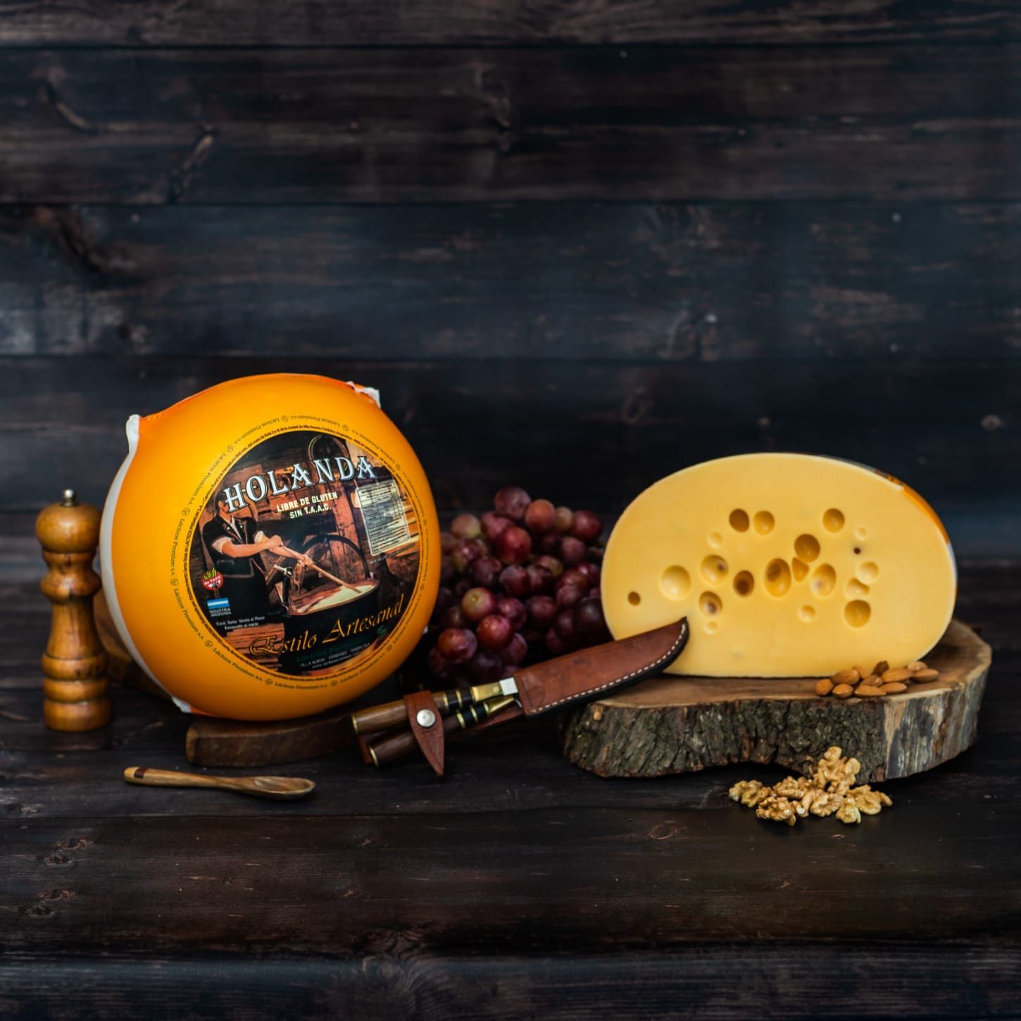 Una rueda de queso holandés con una cuña parcialmente cortada, uvas, un cuchillo y nueces sobre una superficie de madera.
