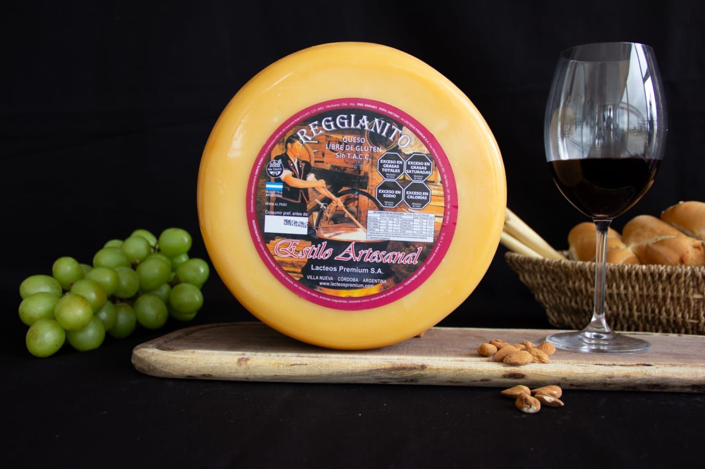 Queso amarillo redondo con etiqueta, uvas, vino tinto, pan y almendras sobre una tabla de madera.