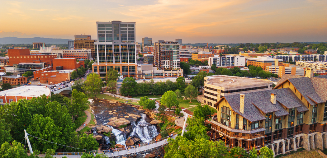 Sunset cityscape in Greenville, South Carolina with modern buildings, trees, and a curved-roof complex by a rocky waterway