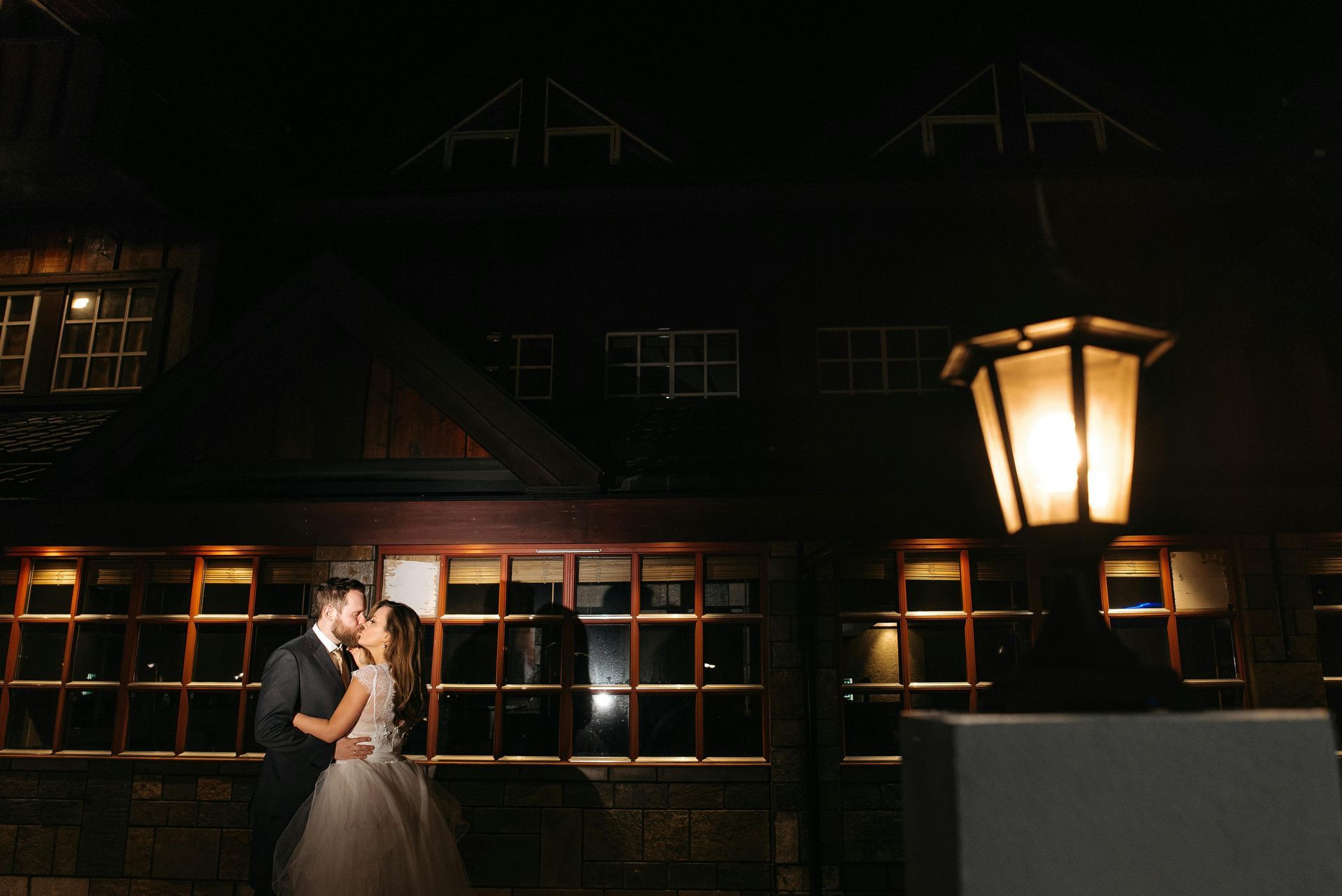 A bride and groom in formal wedding attire embrace outside a rustic building illuminated by a warm lamp at night.