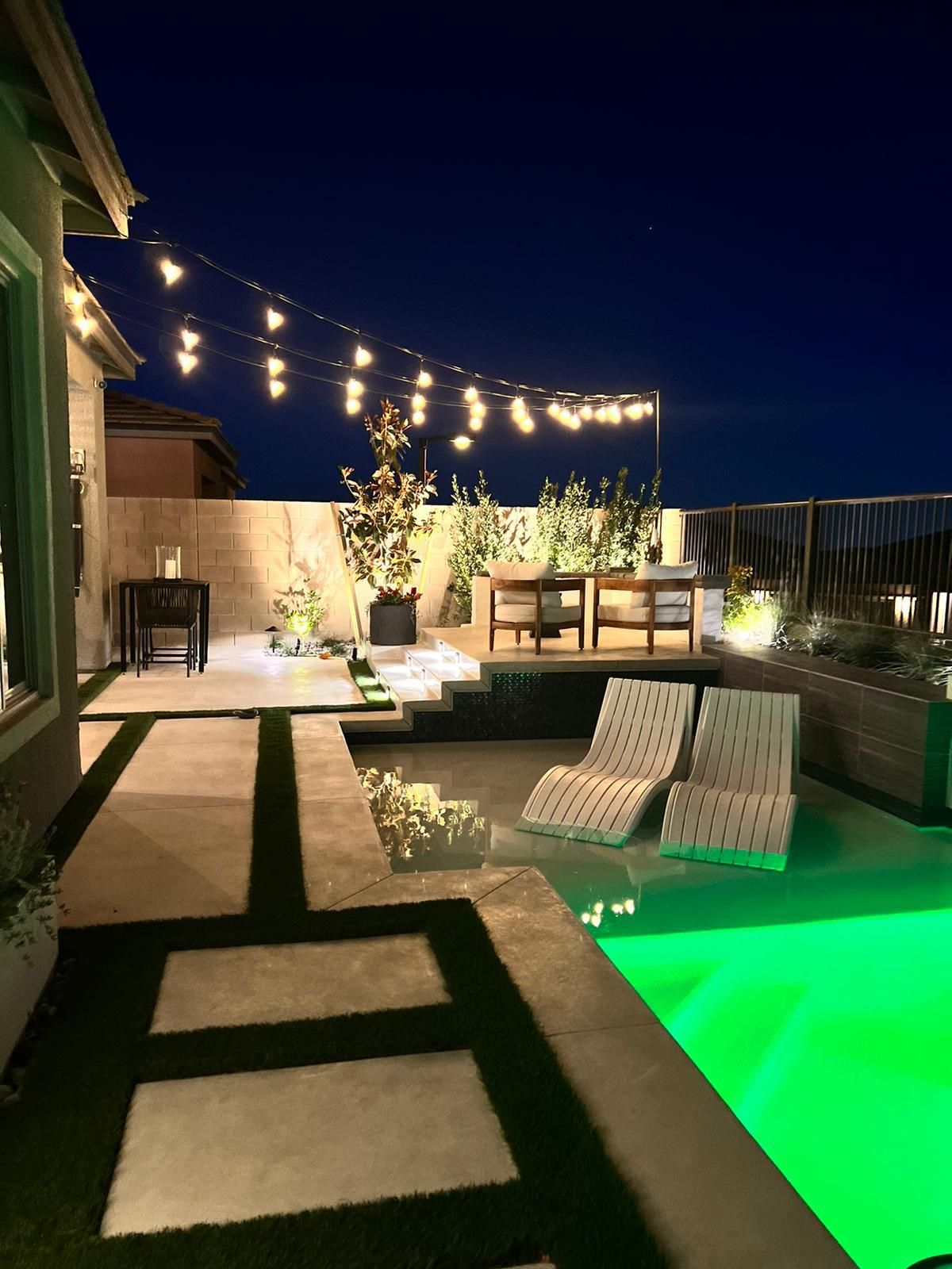 Nighttime pool area with string lights, chairs in pool, green glowing water.