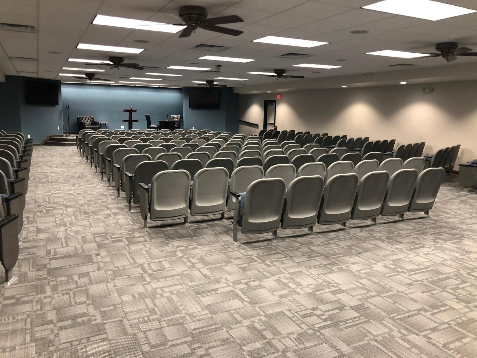 Auditorium with rows of gray seating, ceiling fans, and a podium on a blue wall.