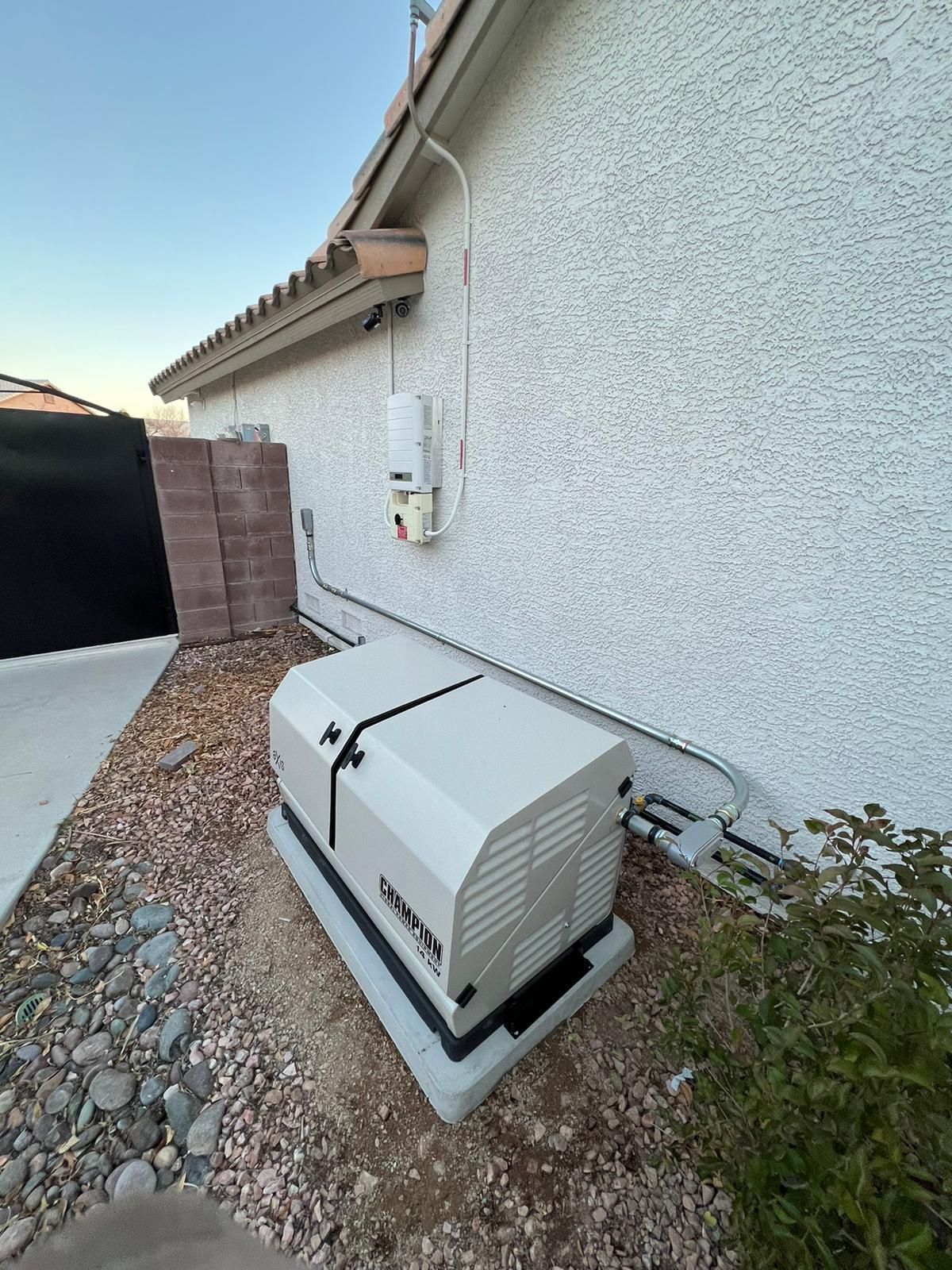 A home generator sits on a concrete pad near a stucco house wall. Gravel, pipes and a power box are visible.