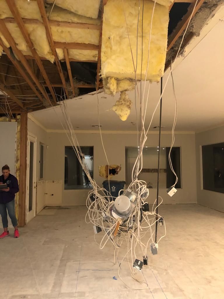 Interior of a house under construction; exposed ceiling with electrical wiring hanging down. Woman stands in the room.