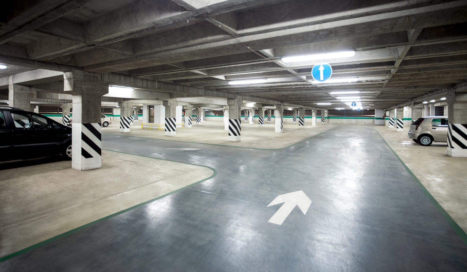 A brightly lit indoor parking garage with concrete columns, painted arrows on the floor, and parked cars.