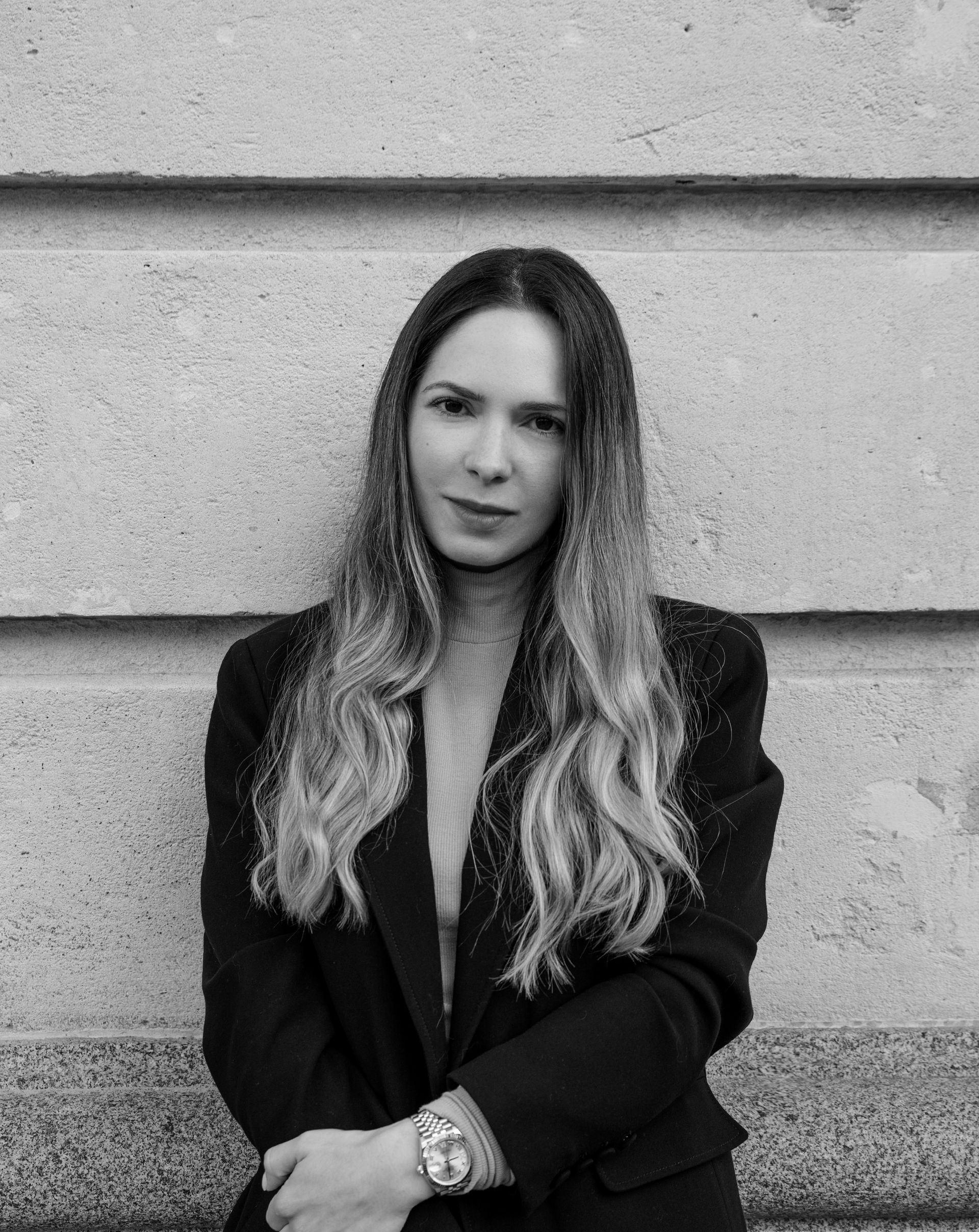 A black and white photo of a woman in a suit standing in front of a stone wall.
