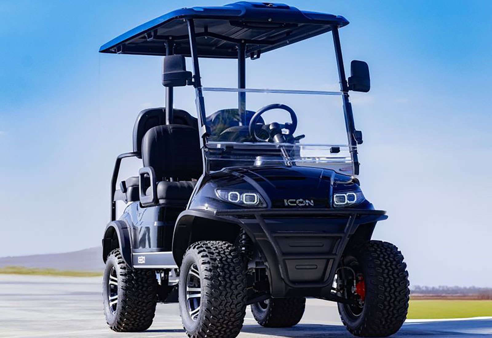Black ICON golf cart with off-road tires, windshield, and roof, on a paved surface against a blue sky.