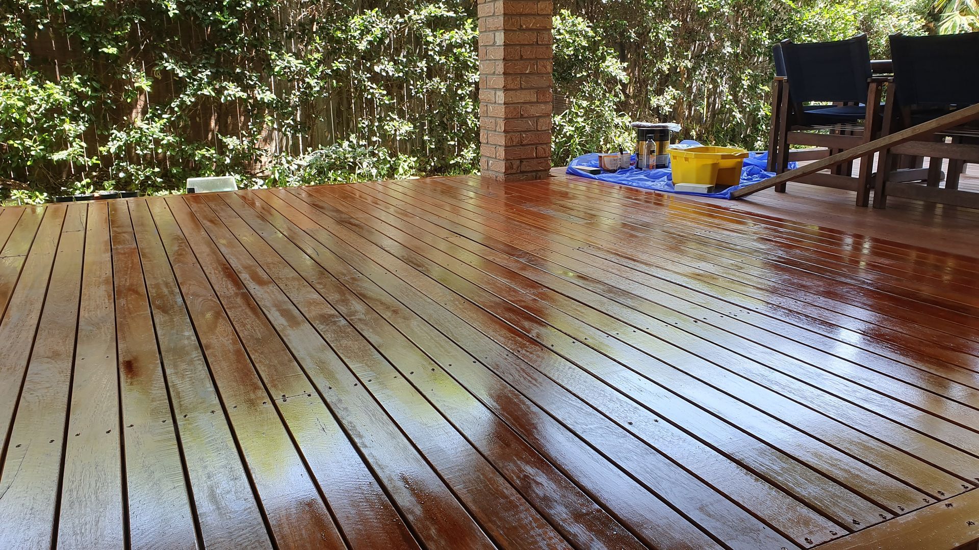 A freshly stained, glossy wooden deck viewed at an angle, with trees in the background and patio furniture on the side.