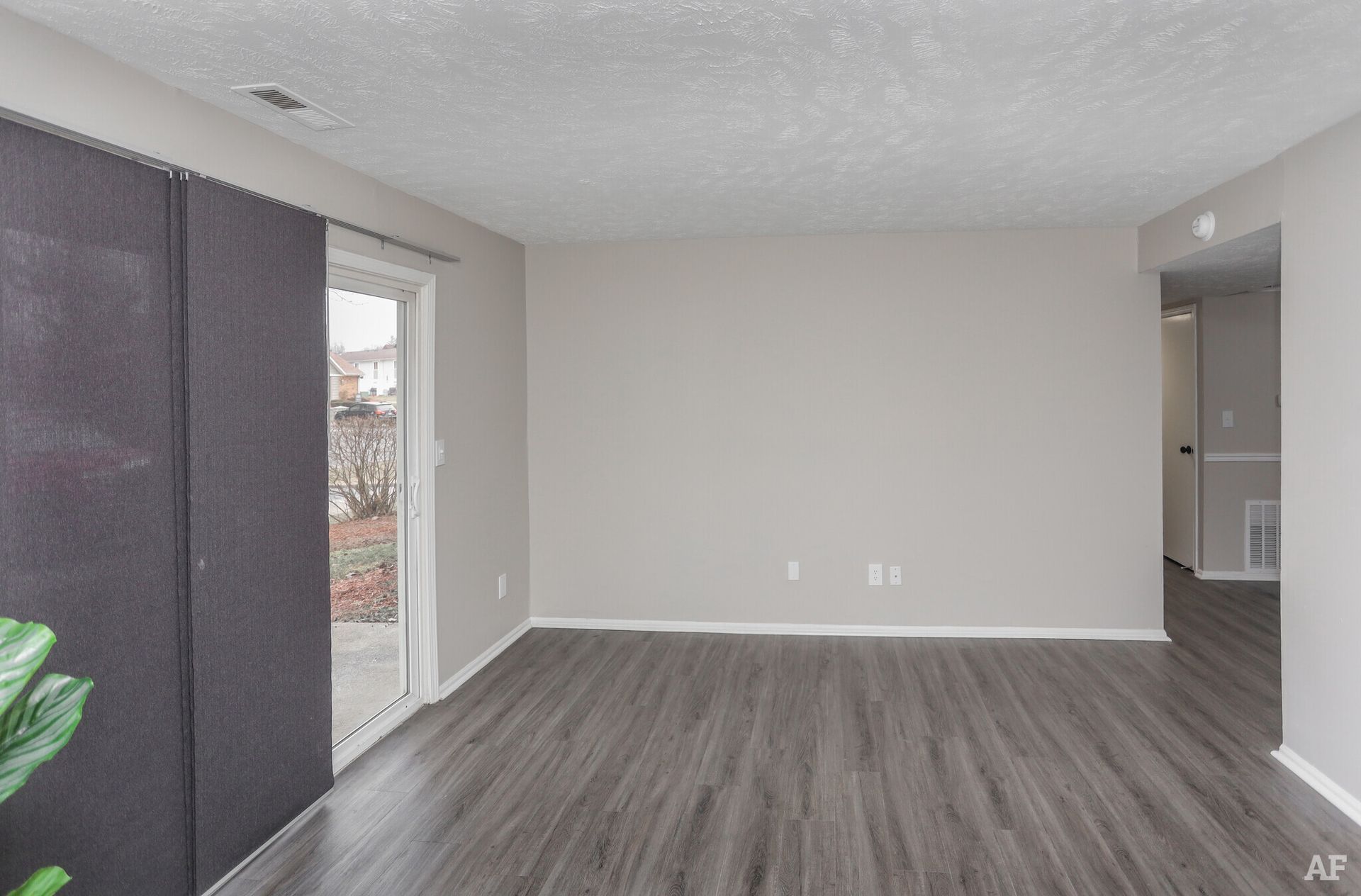 Empty living room with gray walls, dark wood-look flooring, sliding glass door, and entry to another room.