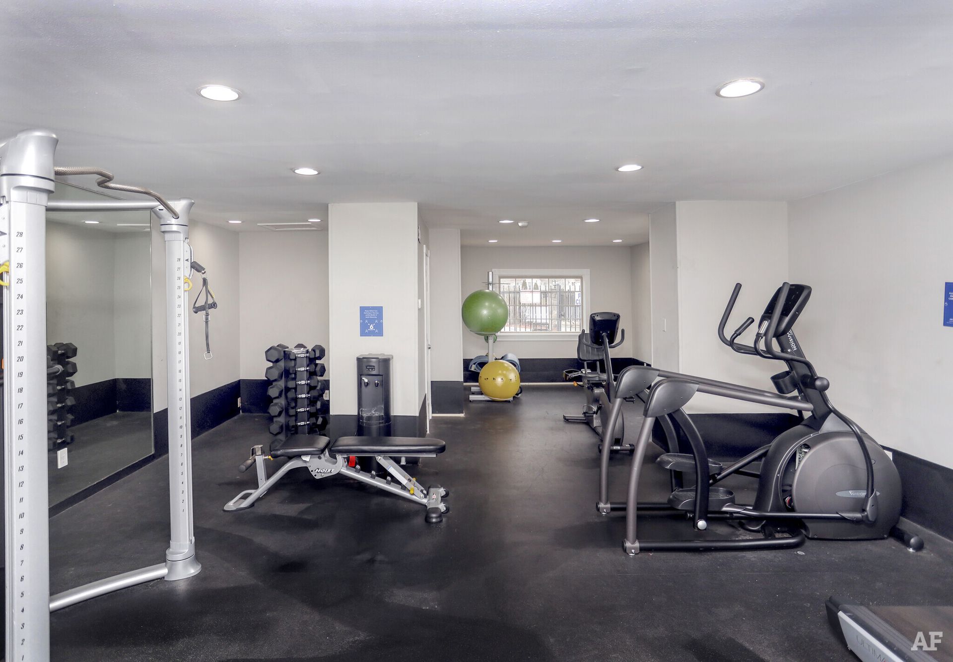 Gym with exercise equipment: weights, elliptical, bench, and exercise balls on a black rubber floor.