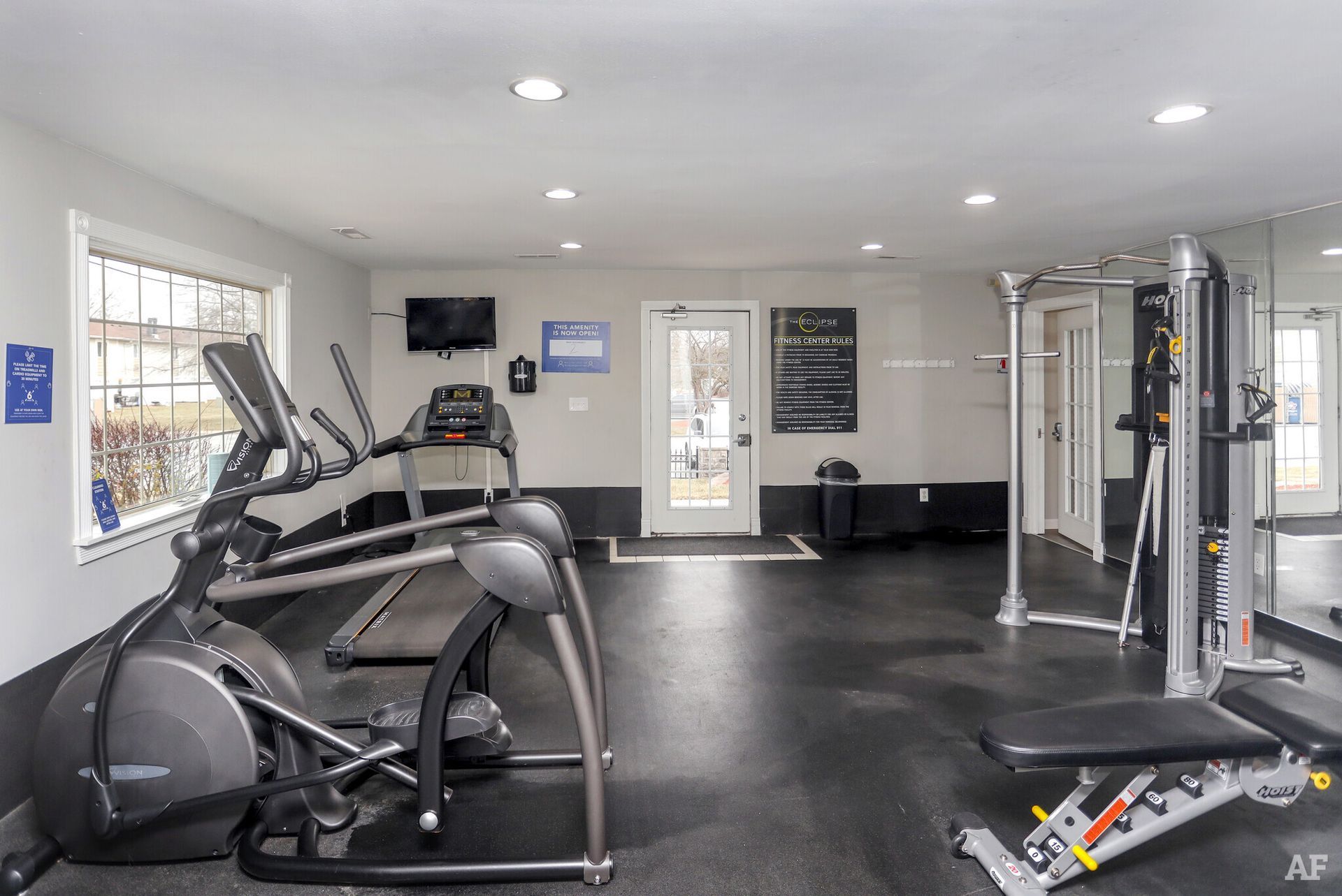 Gym interior with elliptical machine, treadmill, weight machine, bench, TV, and door.