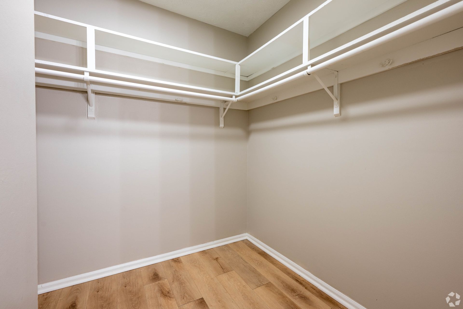 Empty walk-in closet with white shelves and rods, light beige walls, and wood-look flooring.