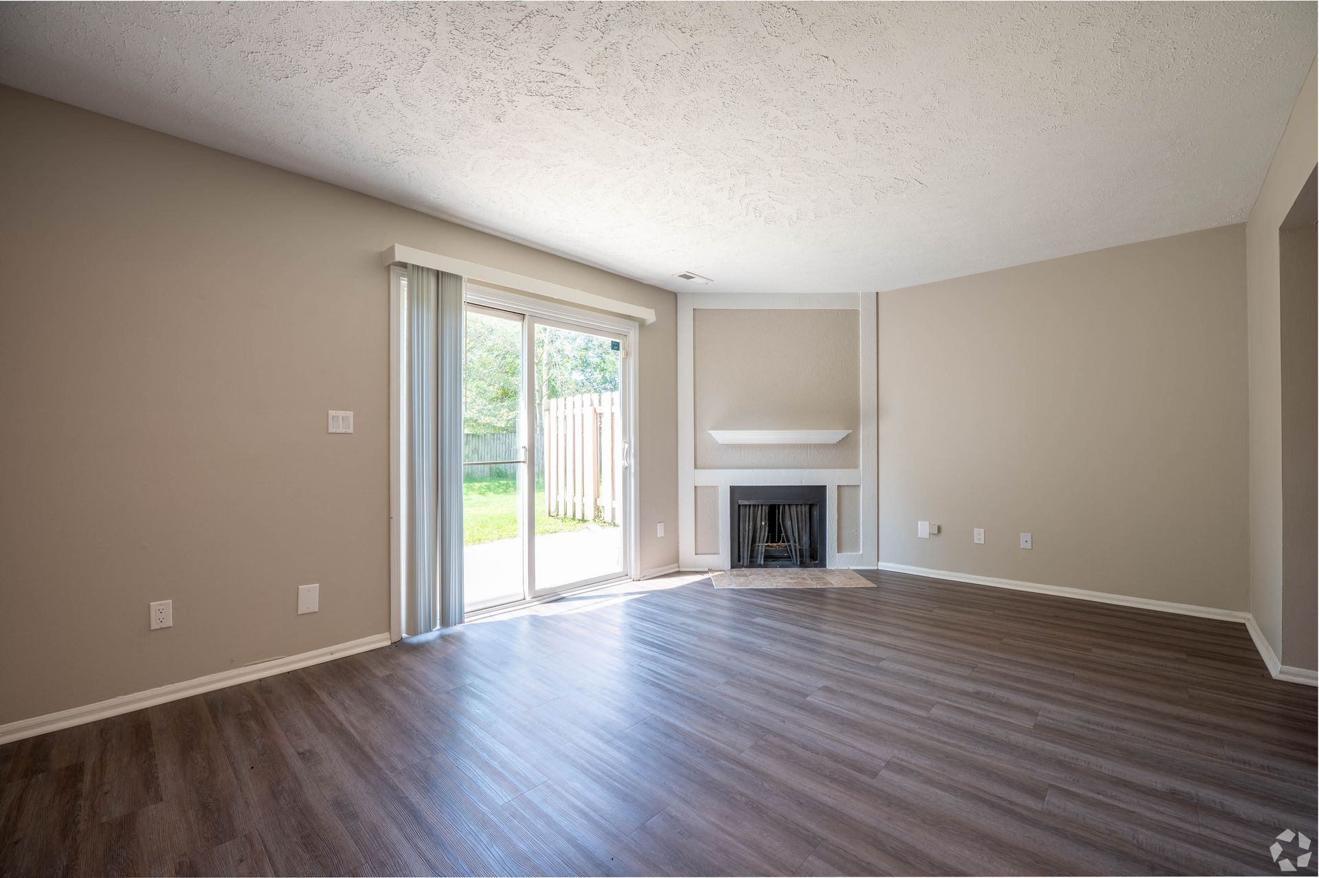 Empty living room with fireplace and sliding glass door; neutral walls, dark wood-look floor.