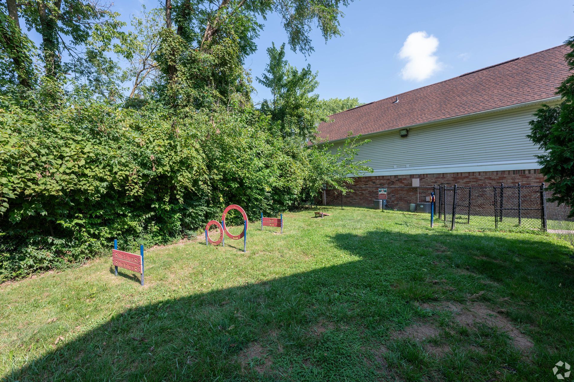 Dog park with agility equipment on grassy ground next to a building with a red roof.