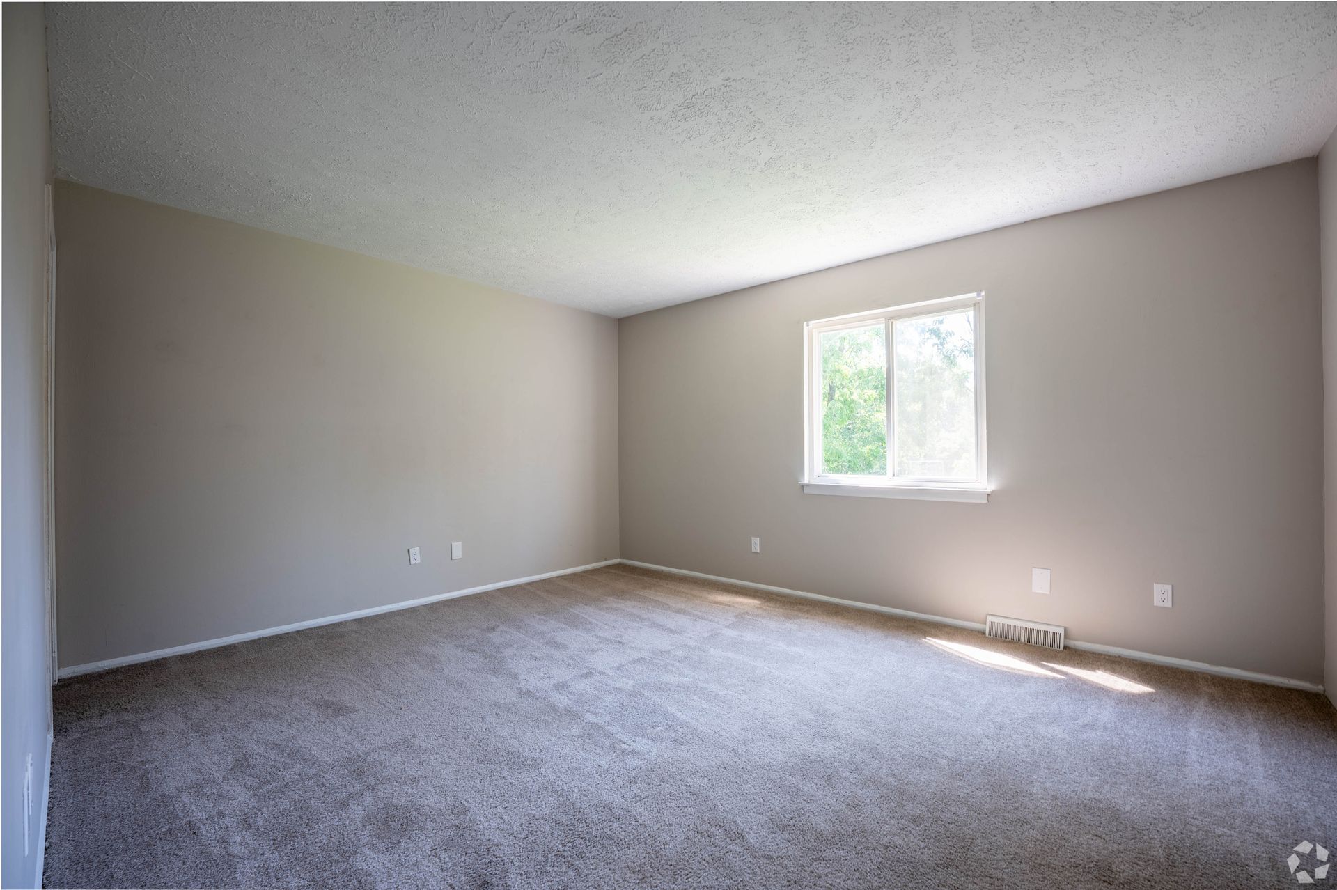 Empty room with beige walls, carpet, and window letting in sunlight.