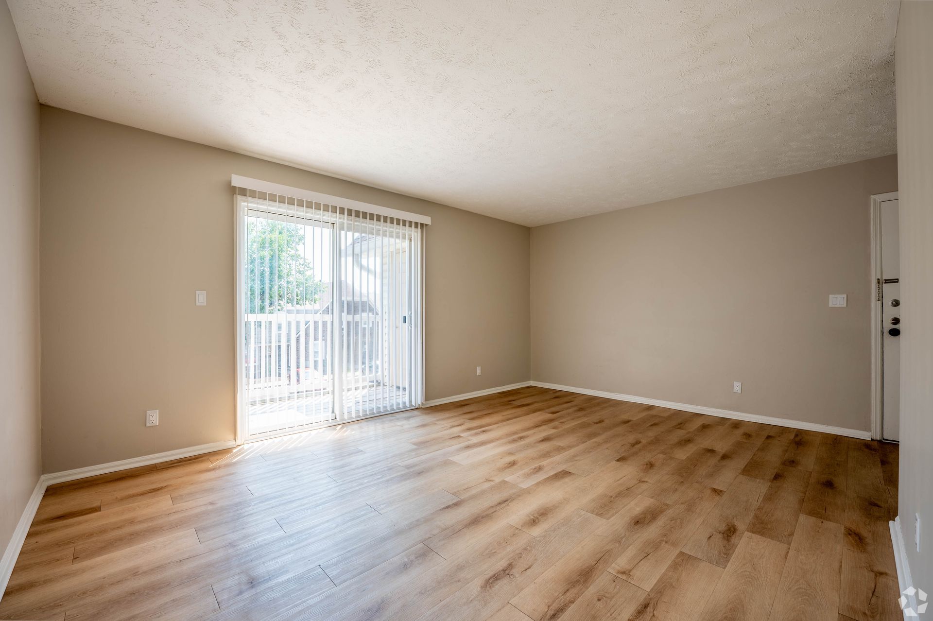 Empty living room with wood floors, tan walls, and a sliding glass door leading to a balcony.