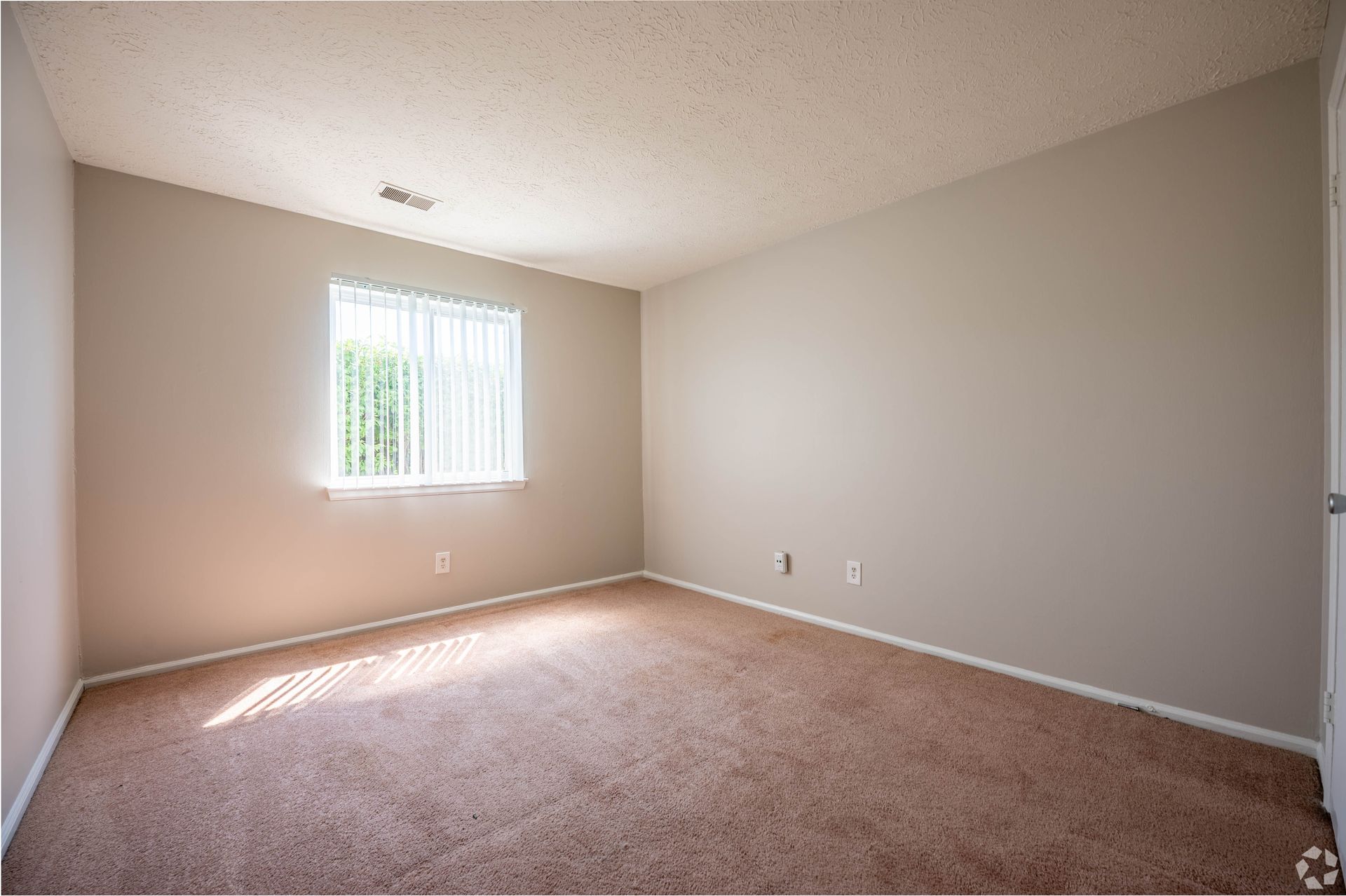 Empty room with beige walls and carpet, a window with blinds, and natural light.