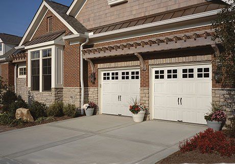 White garage doors with windows, brick and stone house, concrete driveway, with flowers.