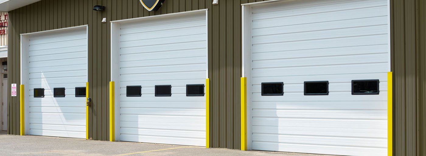 Three white garage doors with black windows, yellow safety posts, and a brown building.