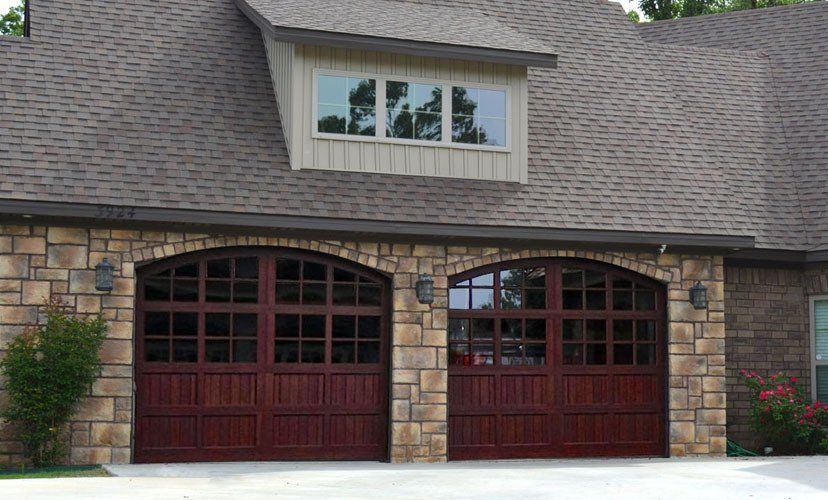 Two mahogany garage doors with arched windows, stone facade, brown roof, and dormer.